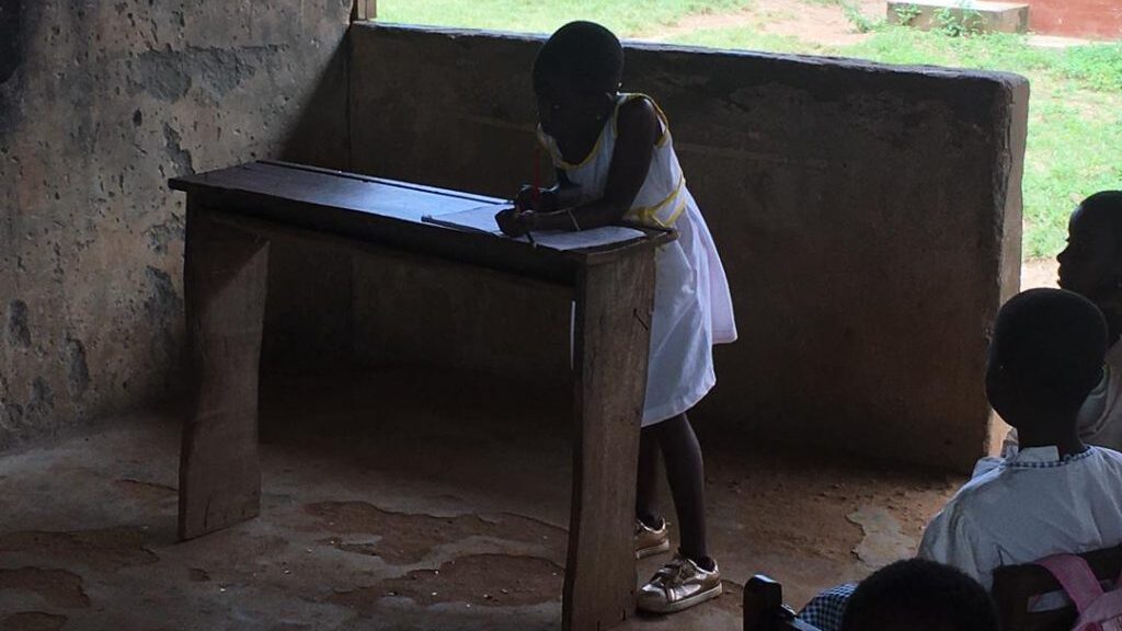 The image shows a young girl writing on a damaged school desk. Supporting the appeal for desks for Adonkwanta